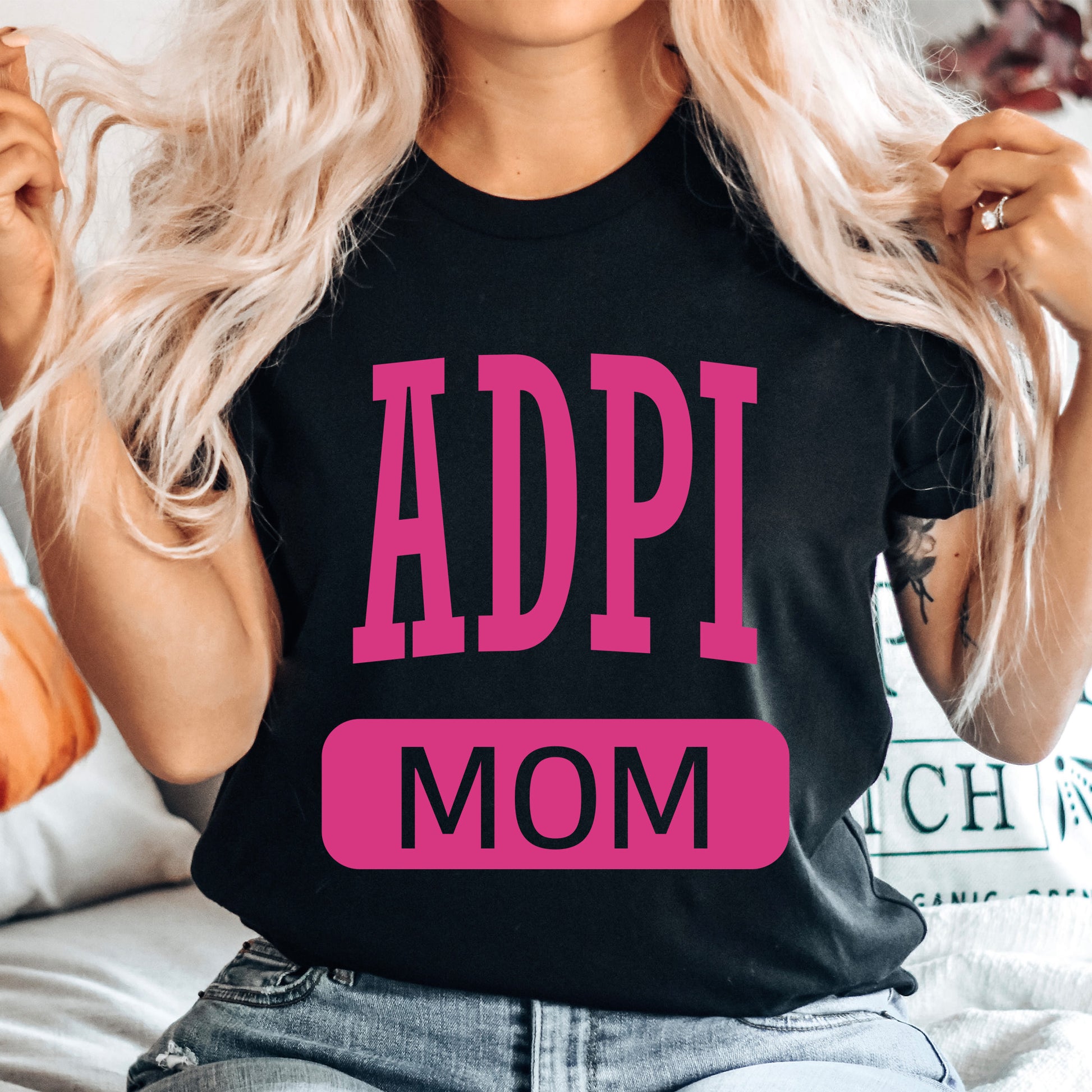 a woman sitting on a bed wearing a t - shirt that says adpi mom