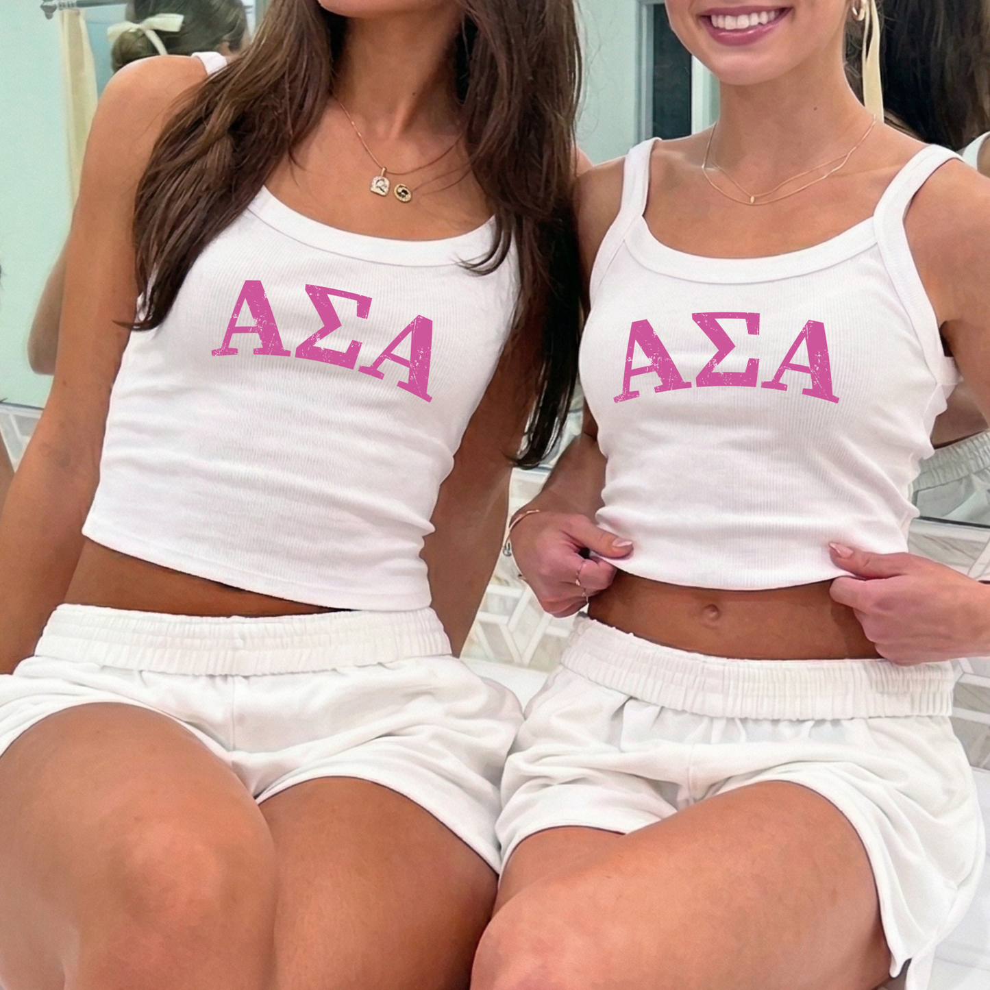 Two women wearing matching white tops with the letters 'ΑΕΛΑ' printed on them.