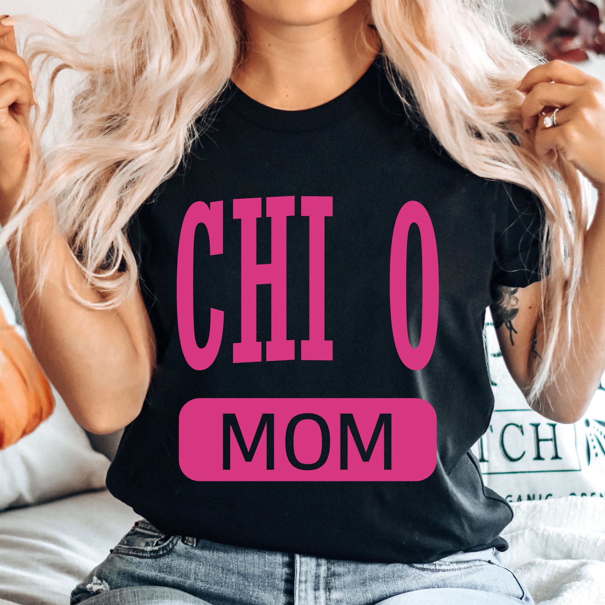 a woman sitting on a bed wearing a black shirt with pink letters