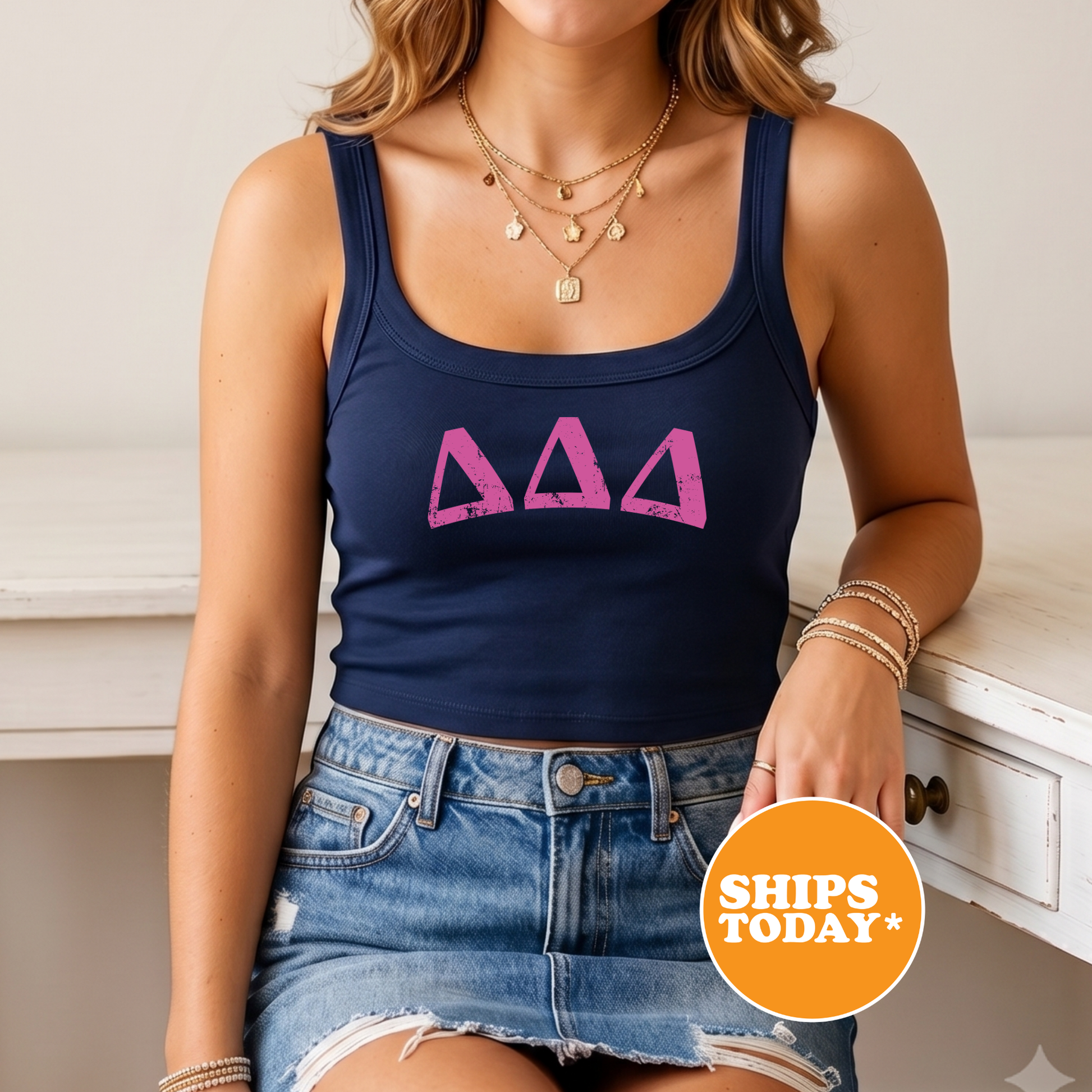 A woman wearing a navy blue crop top with a pink triangle design, denim shorts, and multiple necklaces. She is sitting on a white surface with a "SHIPS TODAY" sign in the foreground.