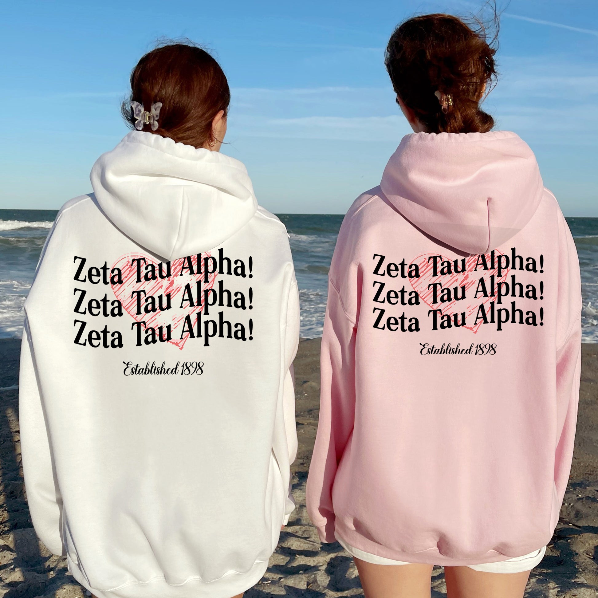 two women standing on a beach wearing matching sweatshirts