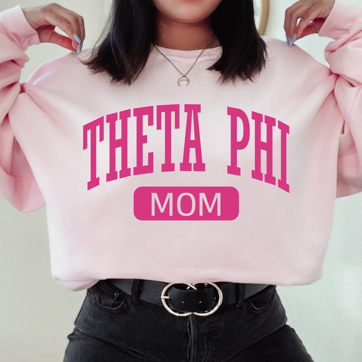 a woman holding a pink sweatshirt with the words theta phi mom on it